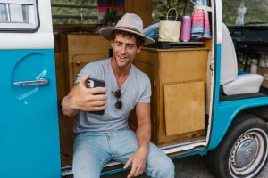 A cheerful young man sits at a campervan door, engaging in a video call, expressing joy and adventure.