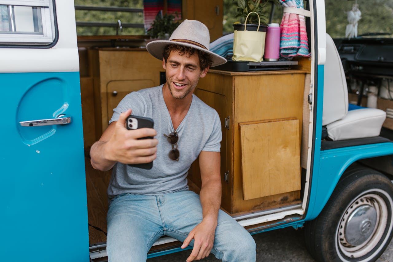 A cheerful young man sits at a campervan door, engaging in a video call, expressing joy and adventure.